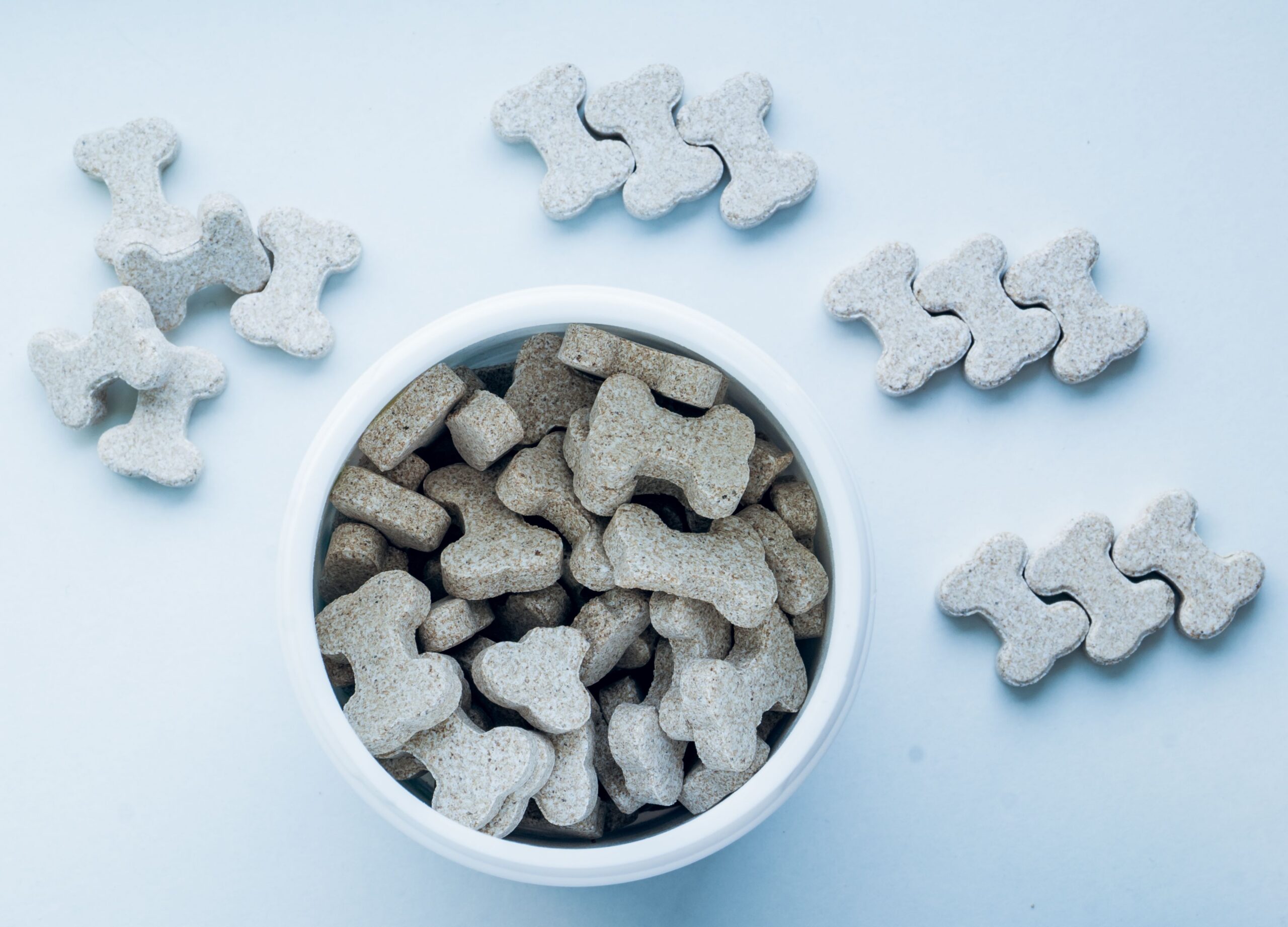 Producten Top view of bone-shaped dog treats in a white bowl, on a light blue background.
