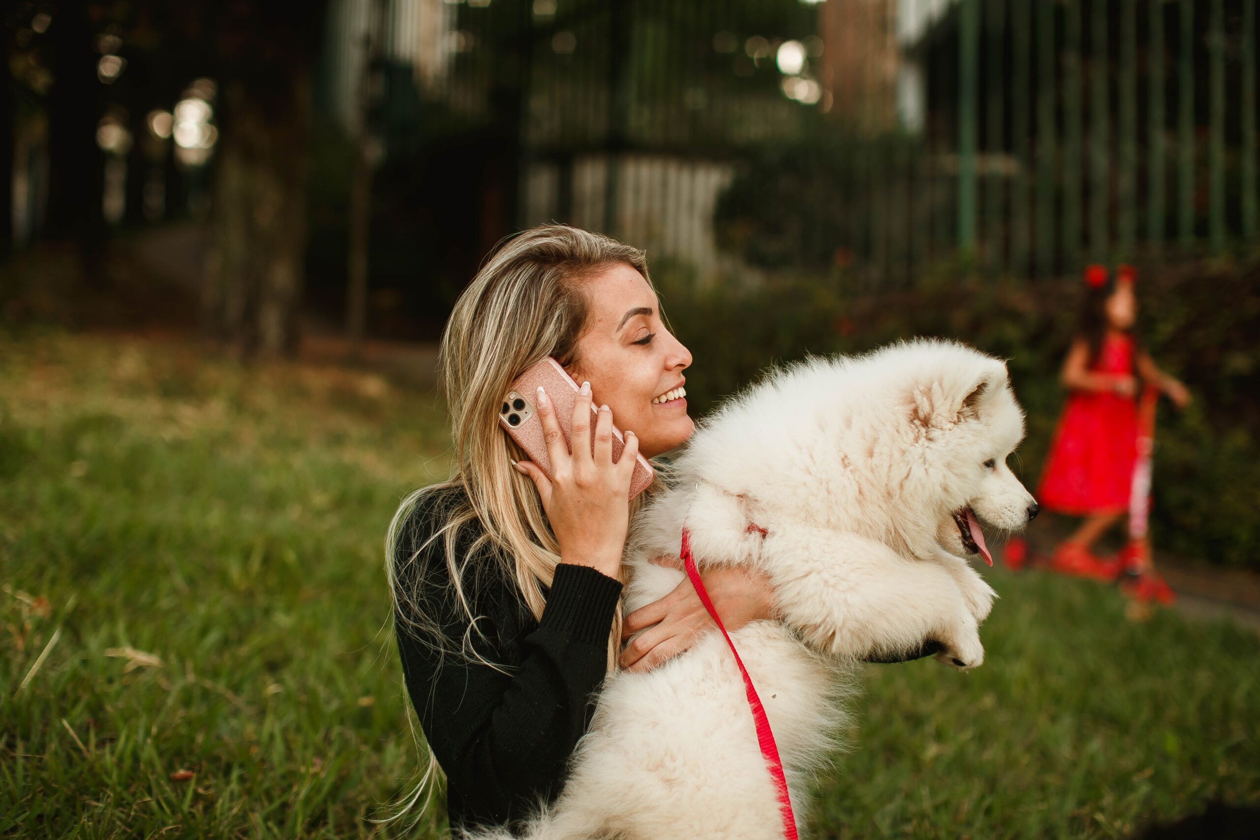 Woman smiling while talking on phone outdoors holding a fluffy white dog.